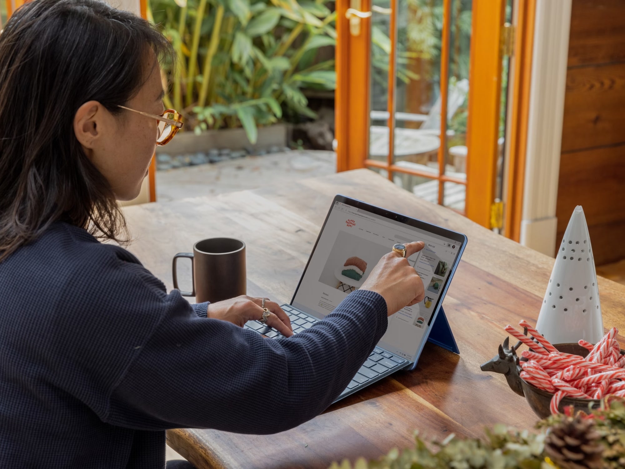 Person wearing glasses using a tablet with keyboard on a wooden table near a cup and a bowl of candy canes.