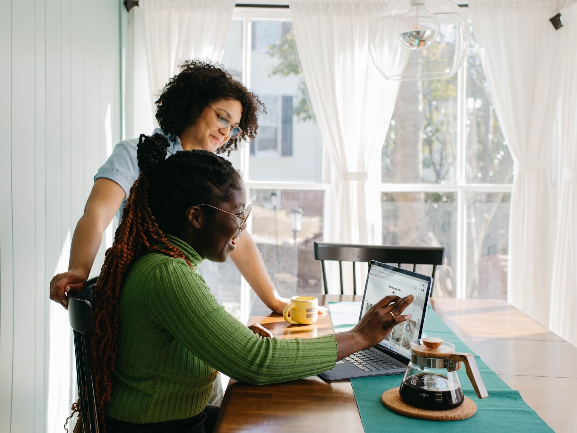 Two women sitting at a wooden table with a laptop and coffee pot, one woman pointing at the laptop screen.