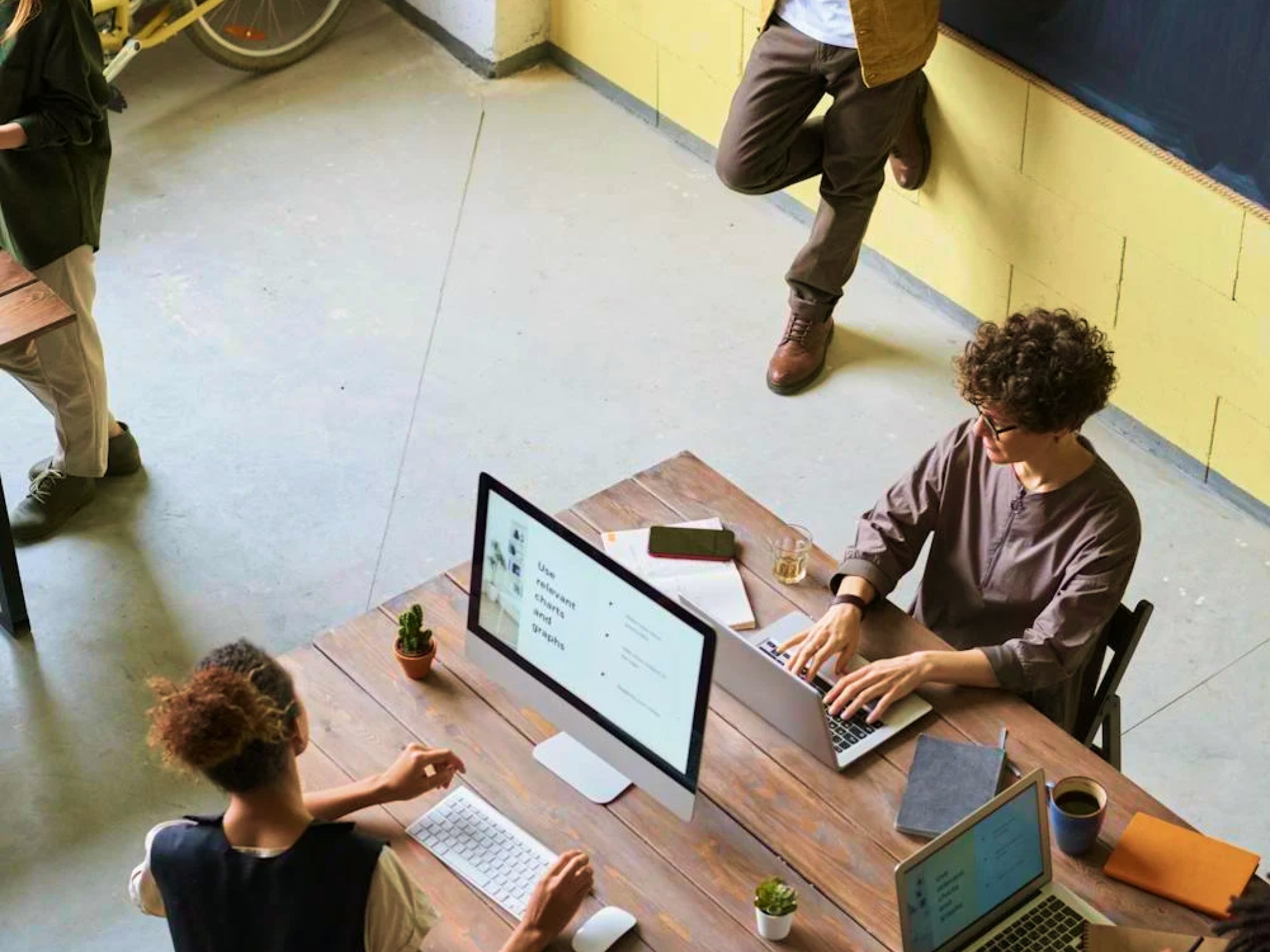 Two people working on laptops at a wooden table in a modern office space with a third person standing nearby.