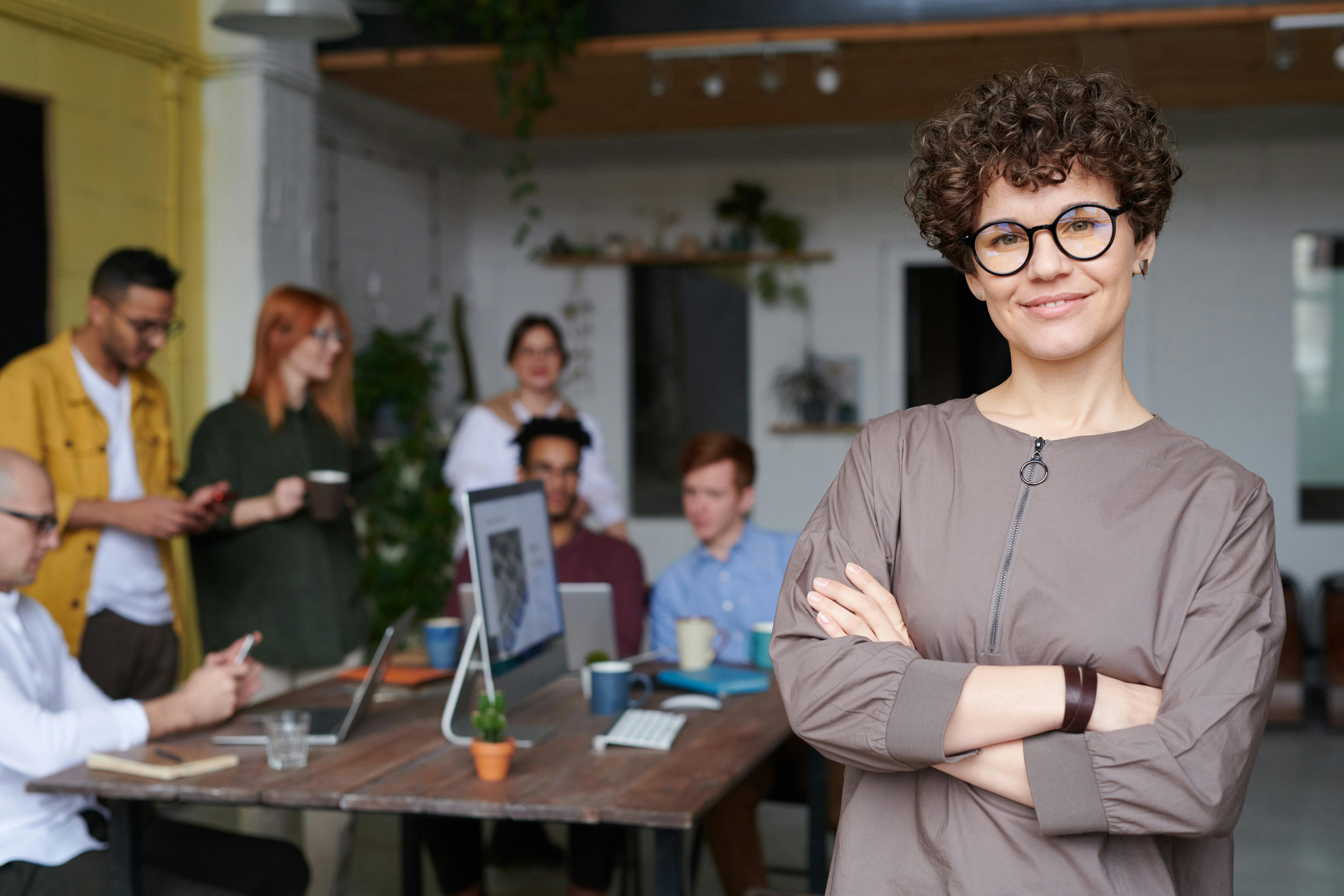 Confident woman with curly hair and glasses standing with arms crossed in front of a group of colleagues working at a wooden table in an office.