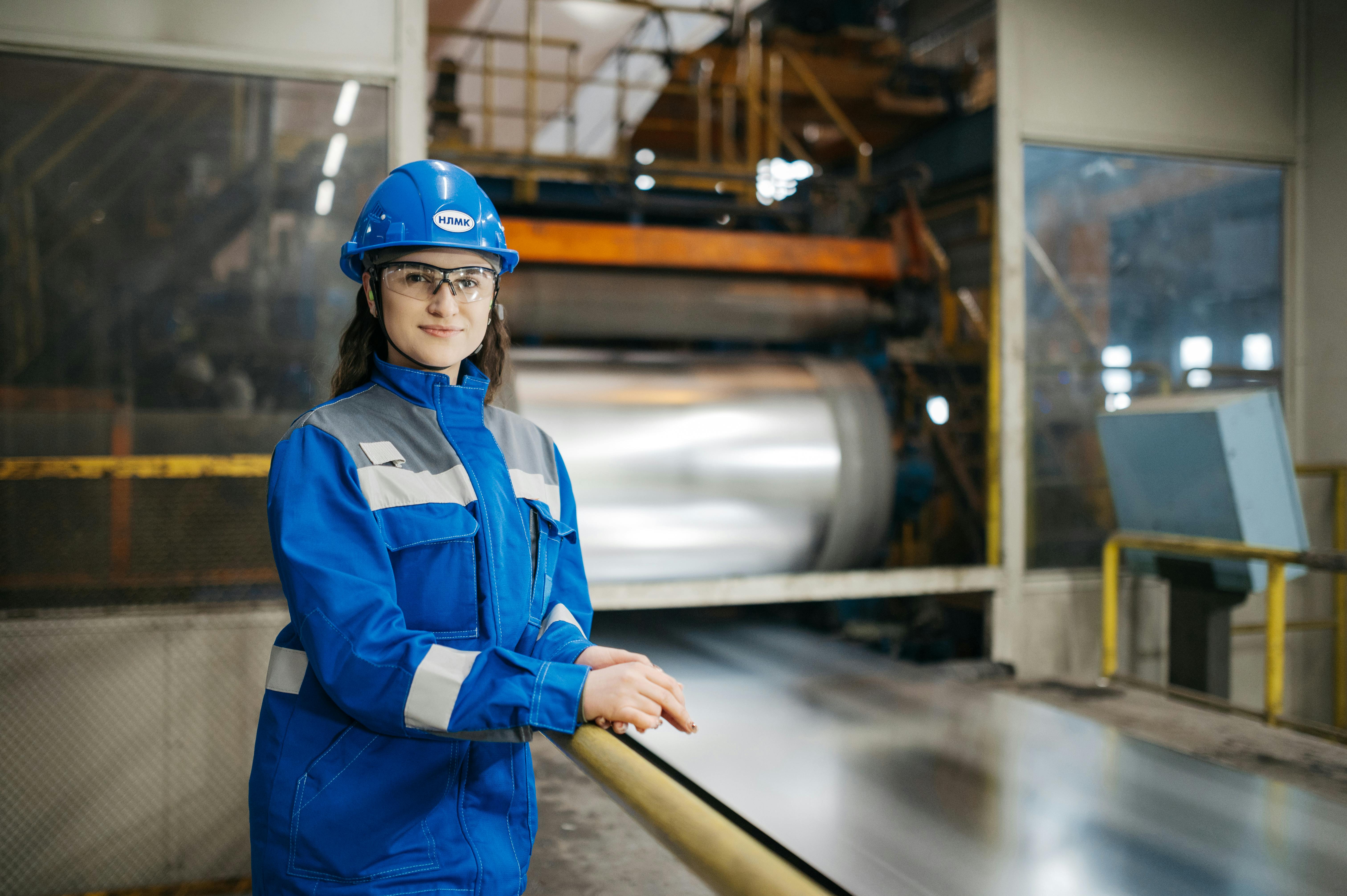 Female factory worker in blue protective gear and helmet standing beside large industrial machinery in a factory setting.