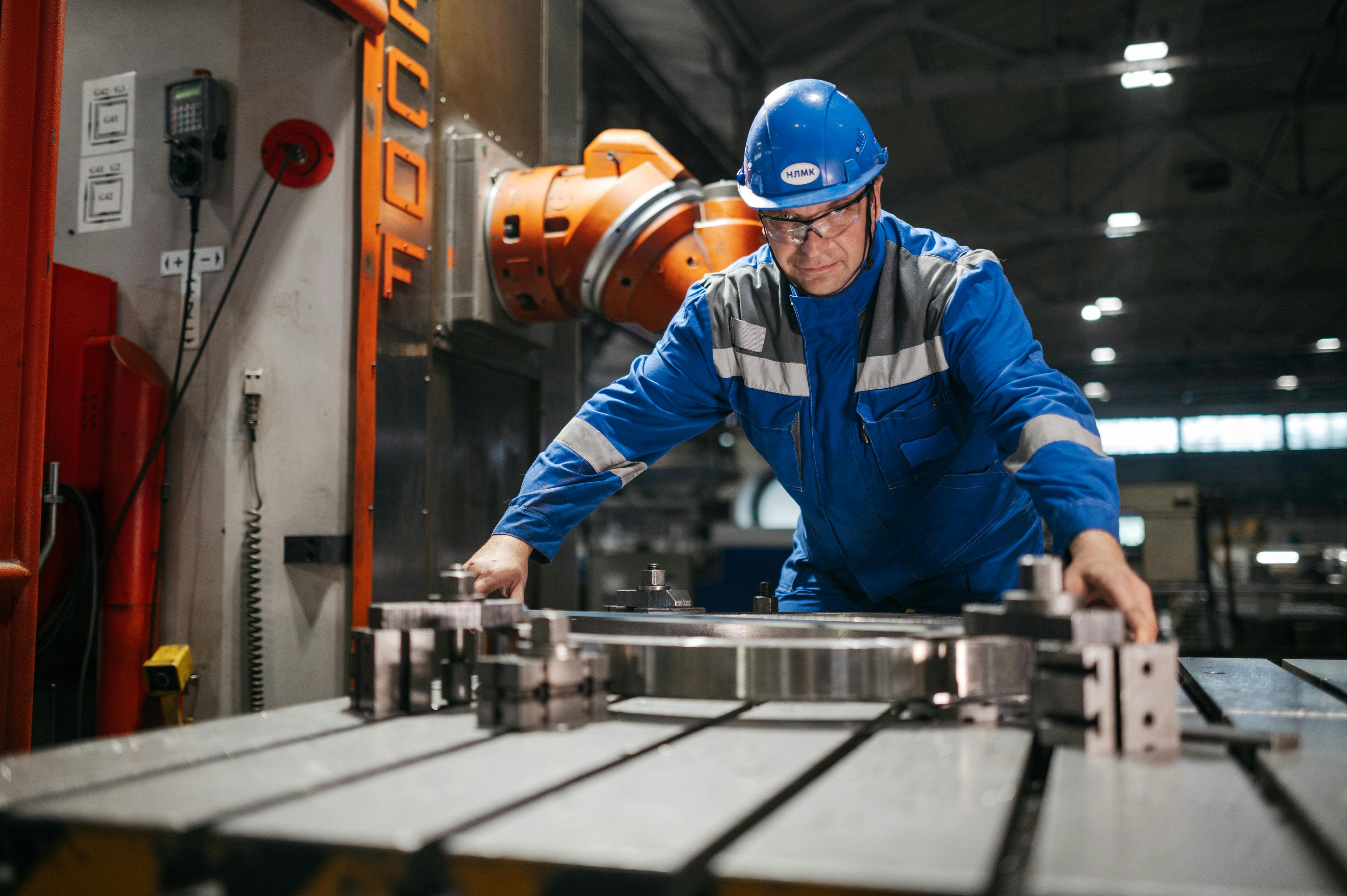 Factory worker in blue protective gear and helmet operating a metalworking machine in an industrial setting.