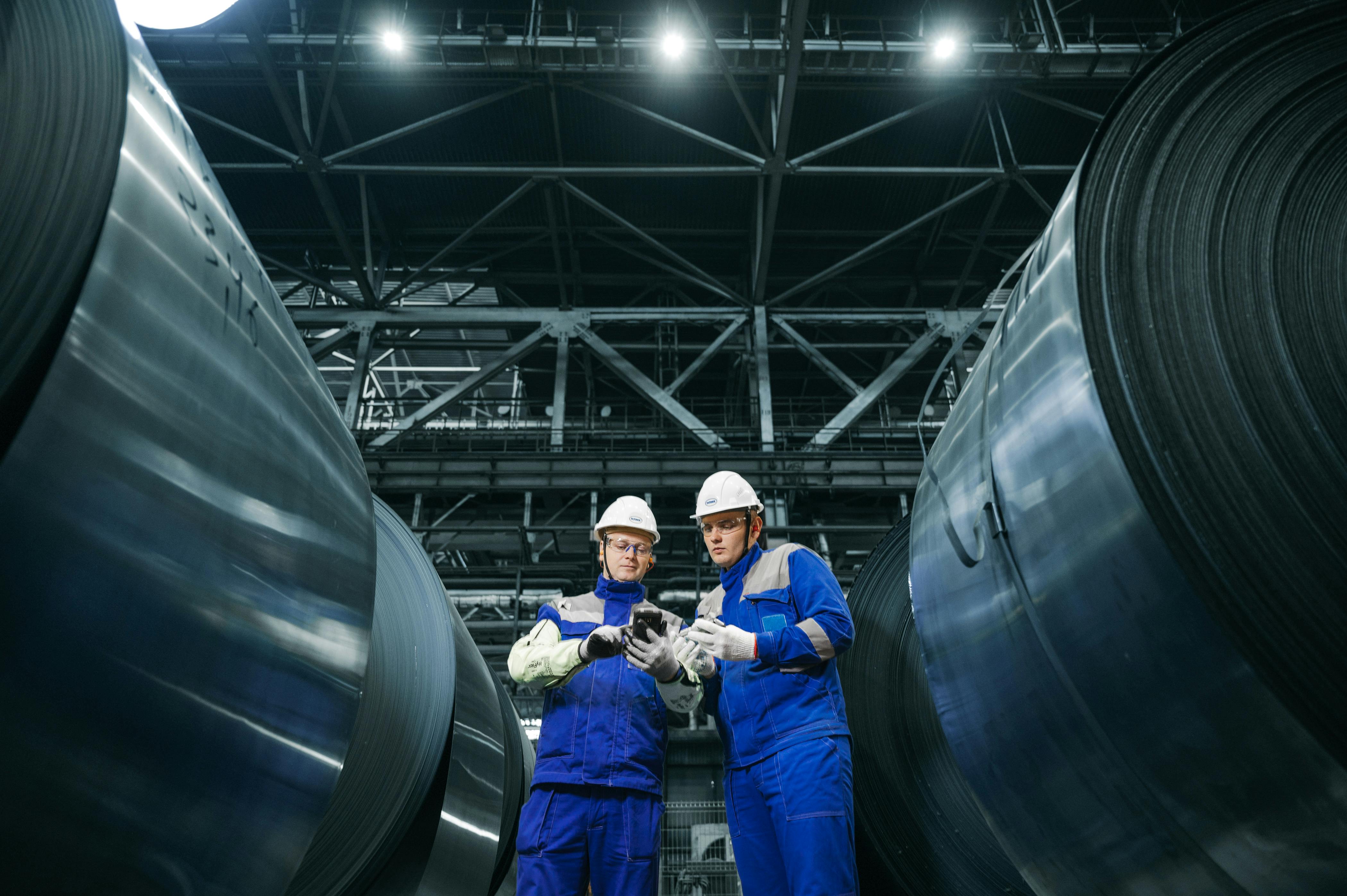 Two factory workers in blue uniforms and white helmets examining a phone between large rolls of metal in an industrial facility.
