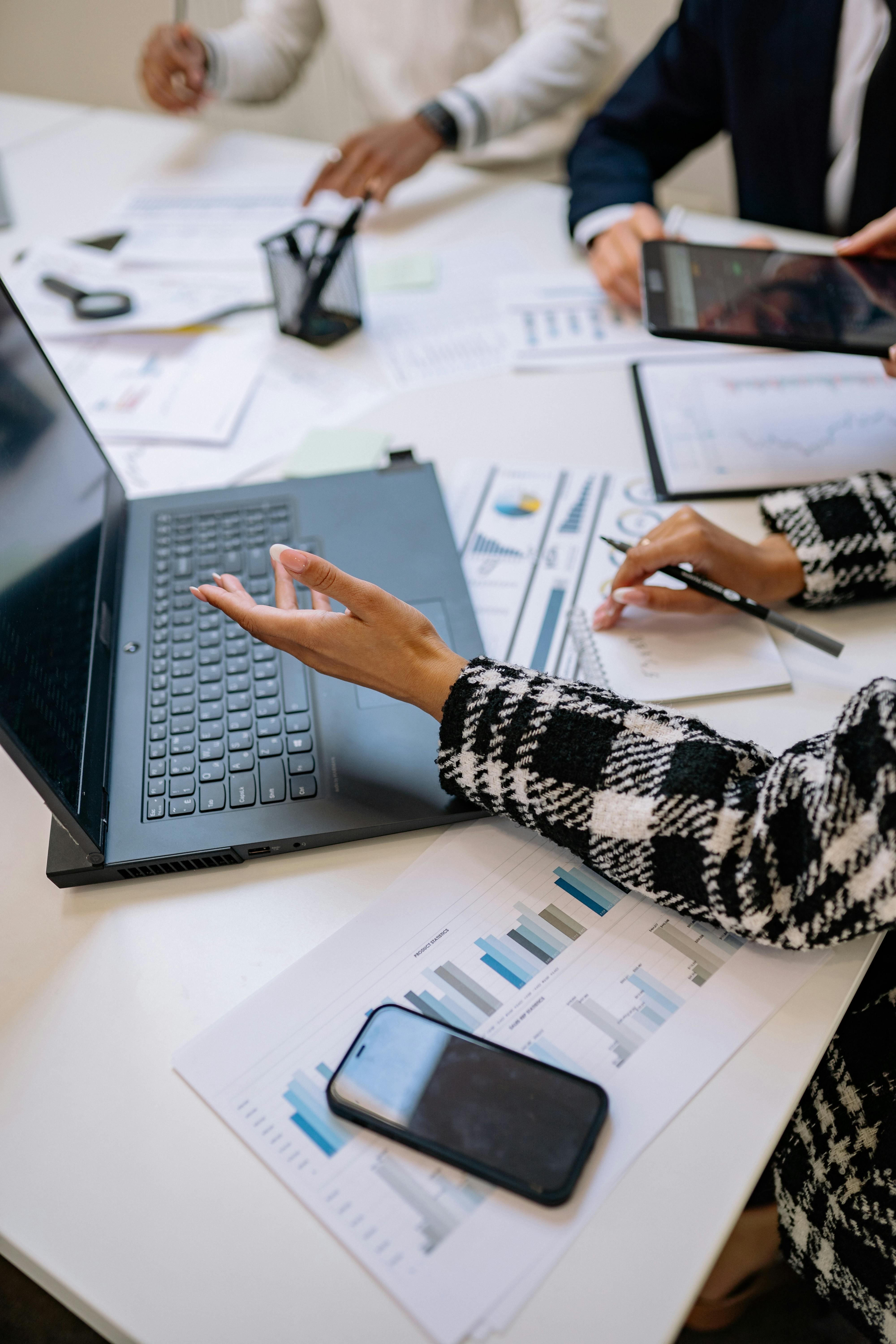 People working at a desk with a laptop, smartphone, papers with charts, and one person writing in a notebook.