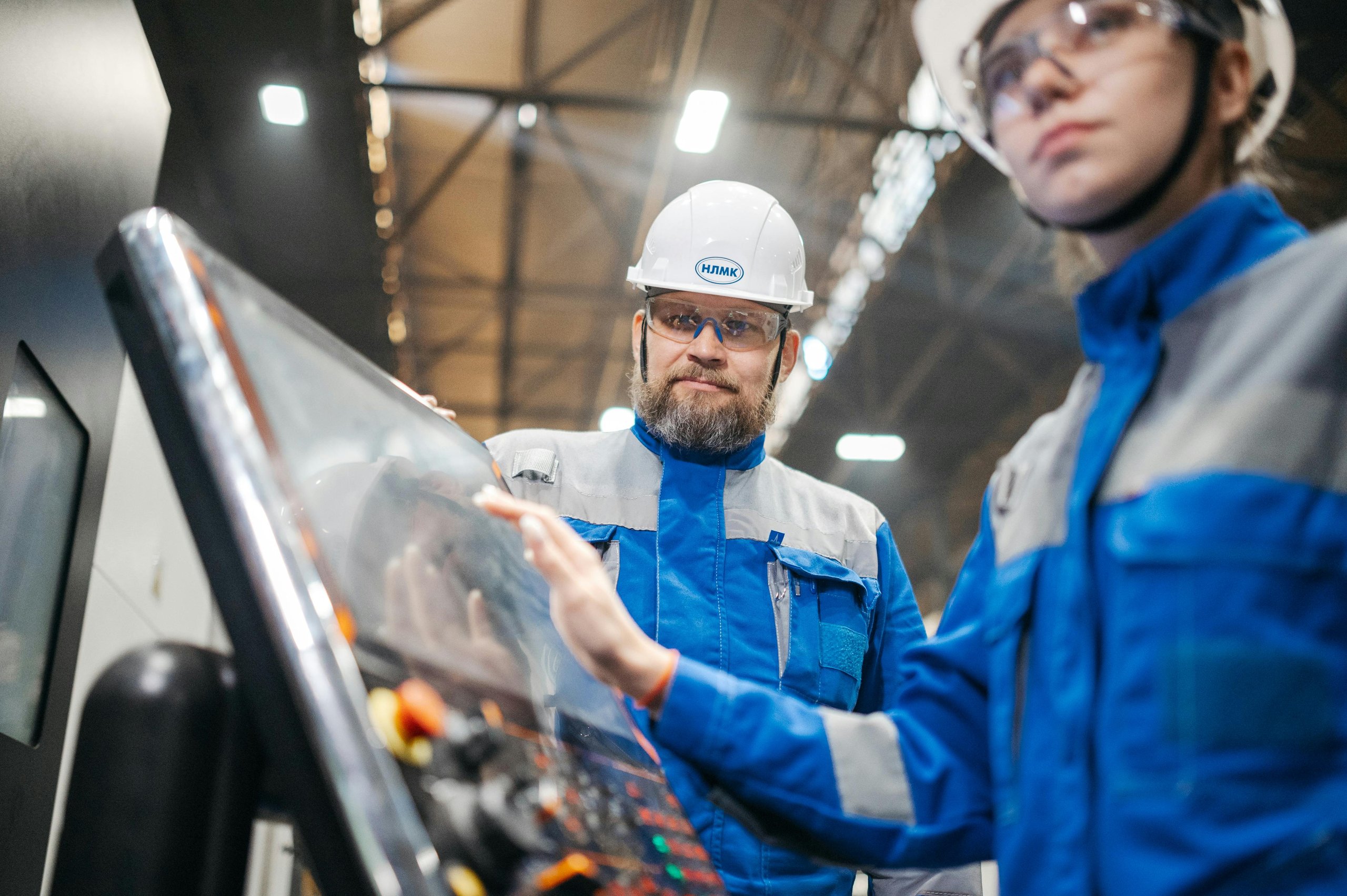 Two factory workers in blue uniforms and white helmets operate a touchscreen control panel inside an industrial facility.