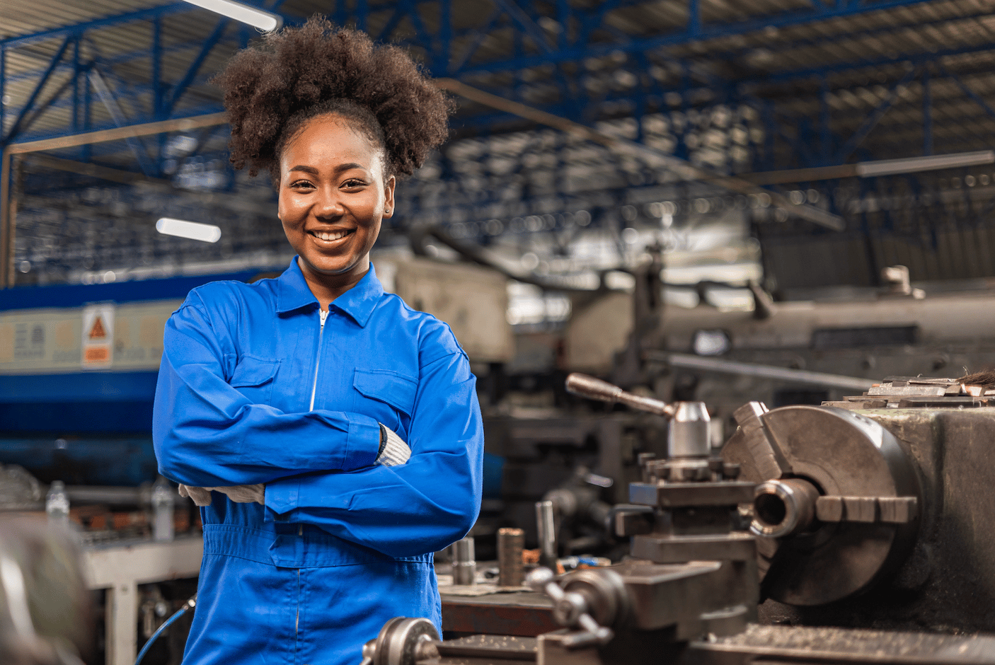 Smiling woman in blue coveralls standing with arms crossed in an industrial workshop.