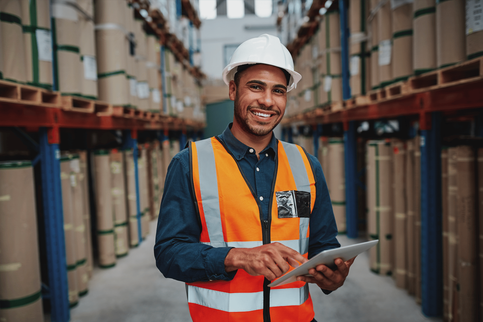 Smiling warehouse worker wearing a white hard hat and orange safety vest using a tablet in an aisle with shelves of stacked packages.