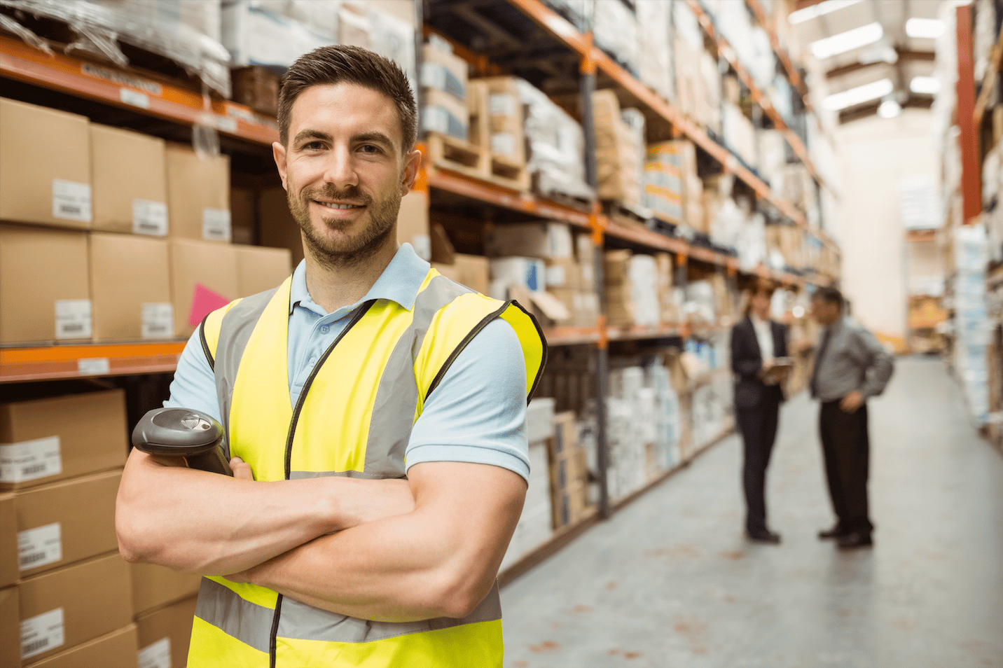 Smiling warehouse worker in a yellow safety vest holding a barcode scanner with shelves of boxes behind him.
