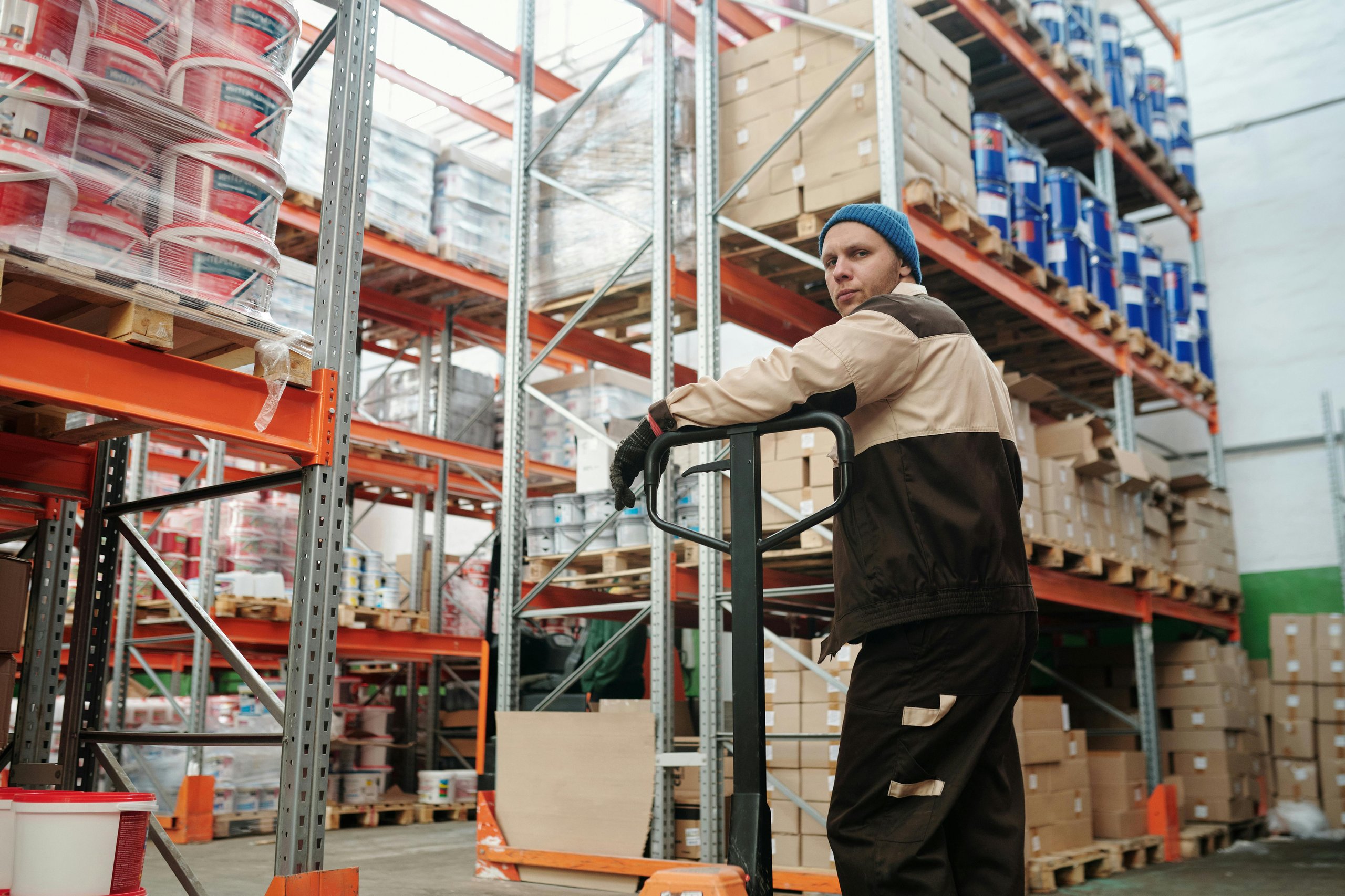 Warehouse worker in uniform and blue beanie operating a pallet jack among shelves stacked with boxes and paint containers.
