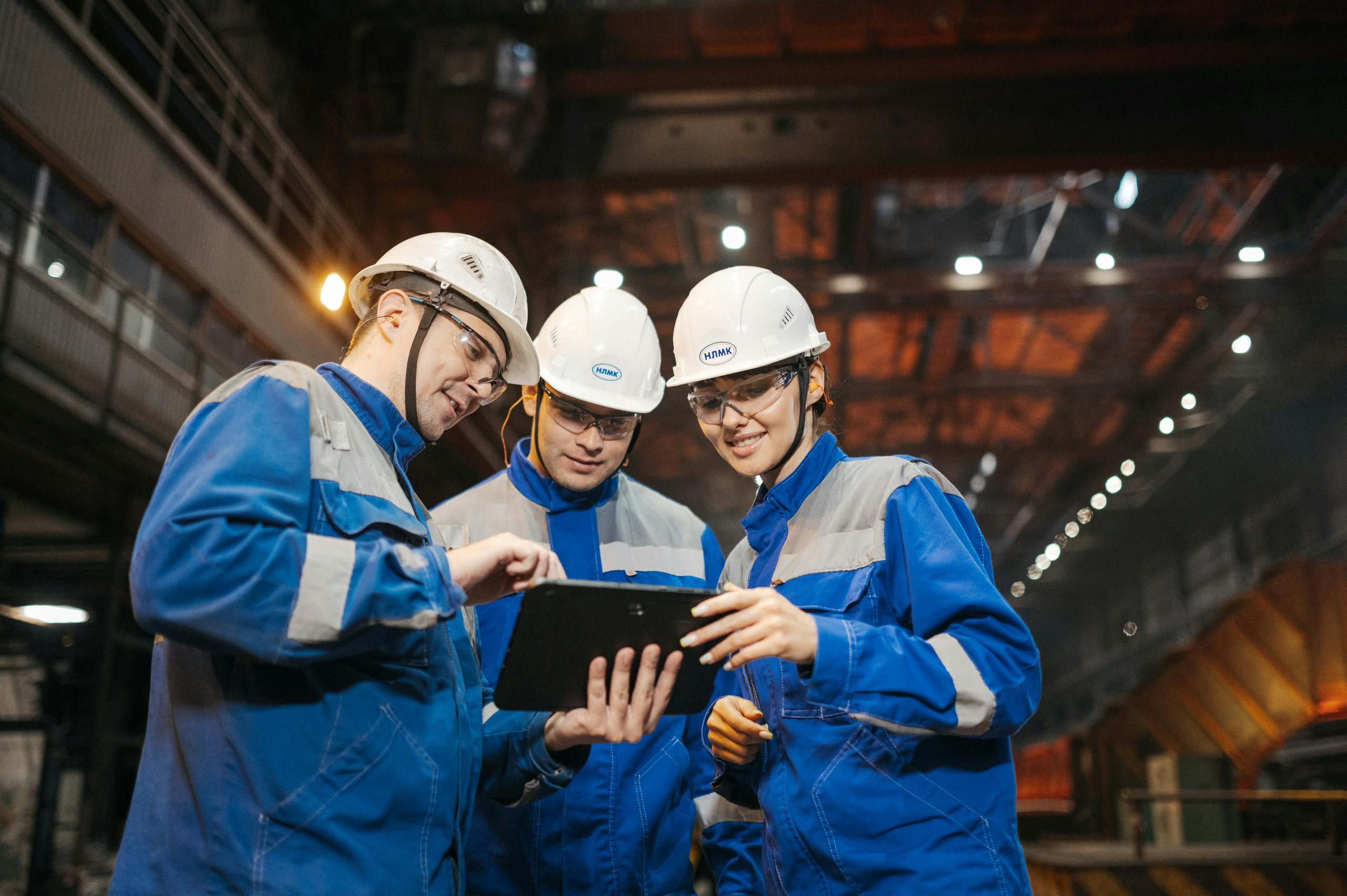 Three factory workers wearing blue uniforms and white helmets reviewing information on a digital tablet inside an industrial facility.