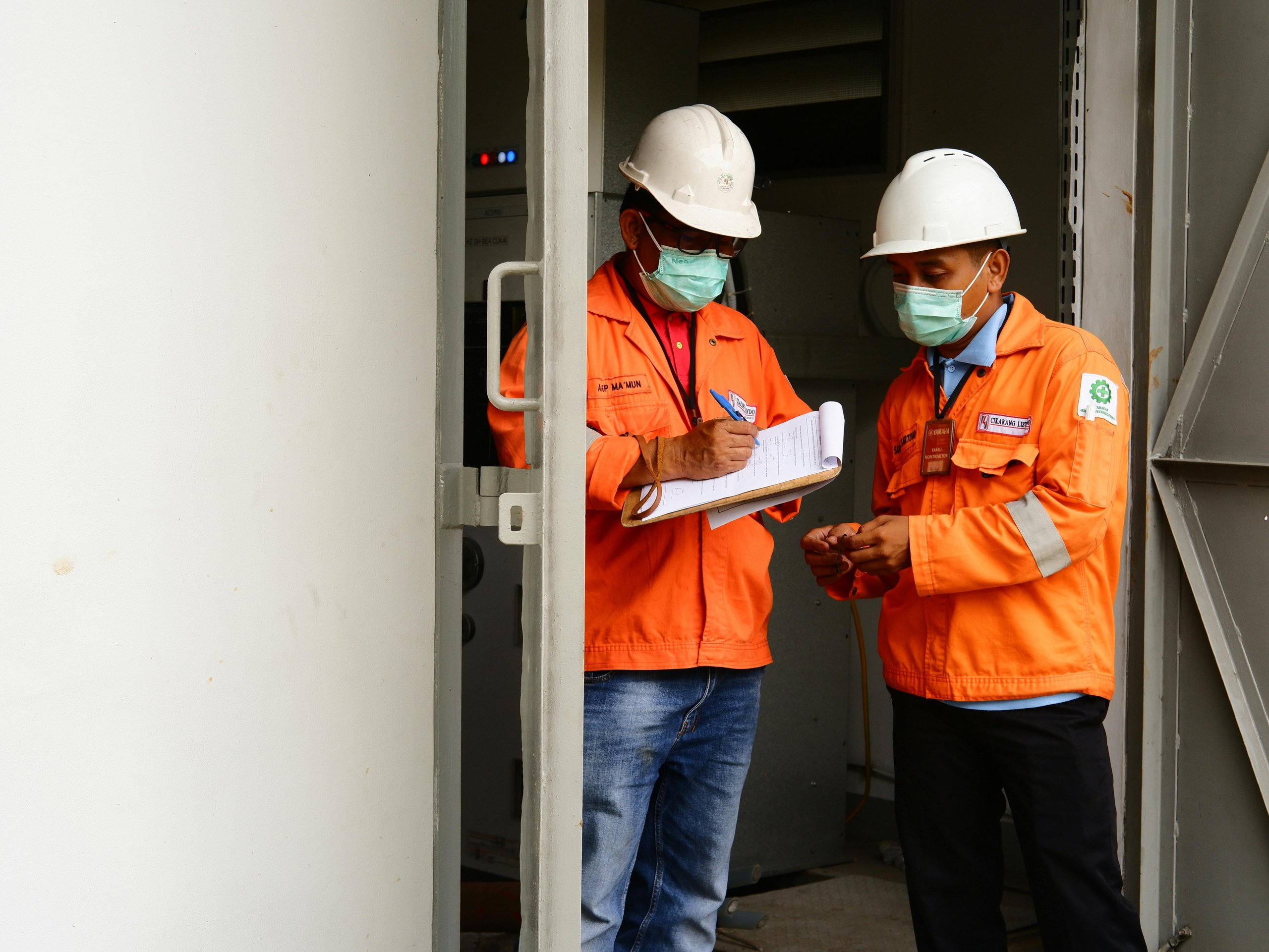 Two construction workers wearing orange jackets, white helmets, and face masks, reviewing documents inside an industrial setting.