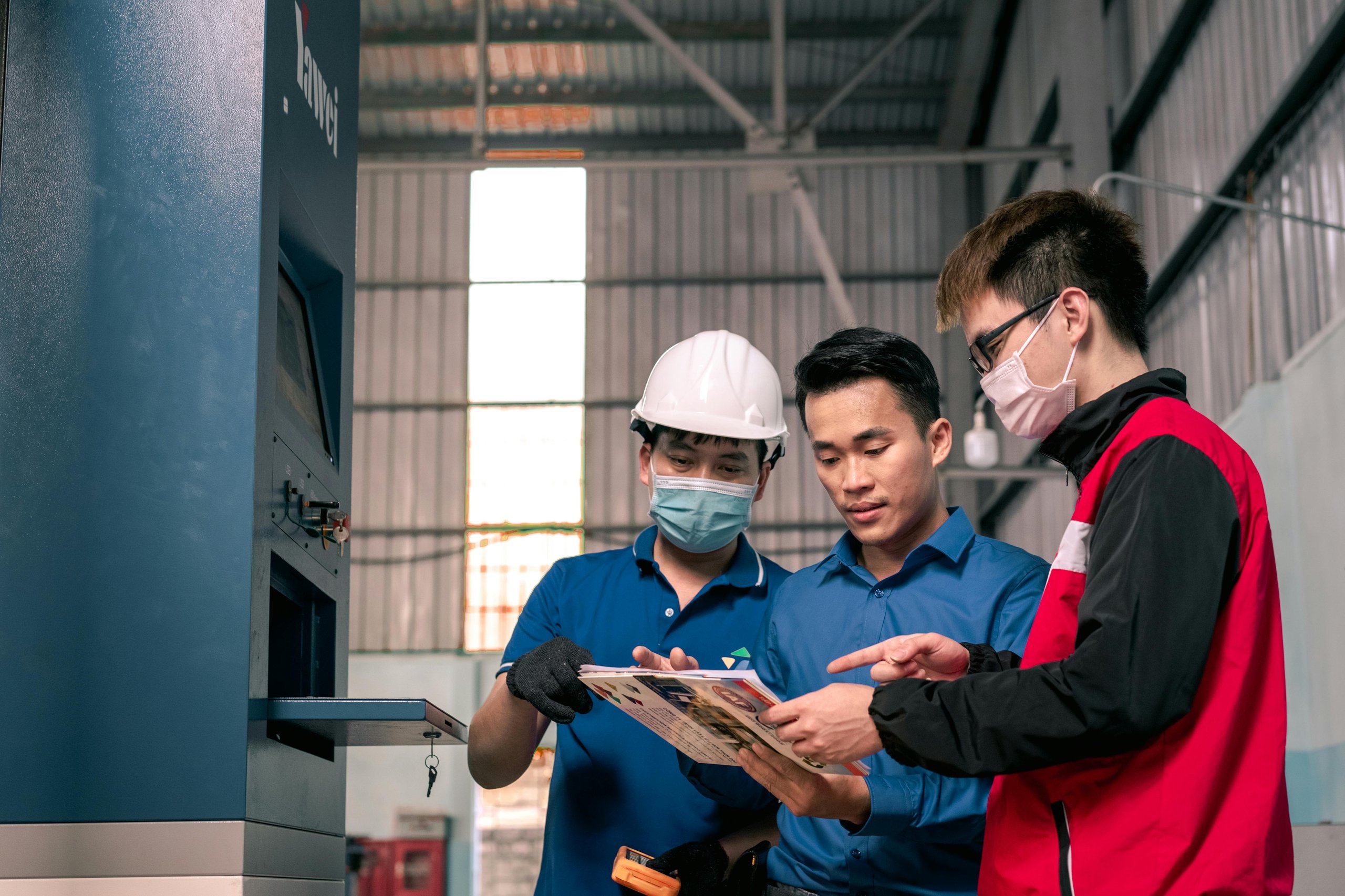 Three men in a workshop discussing documents, one wearing a white hard hat and face mask.