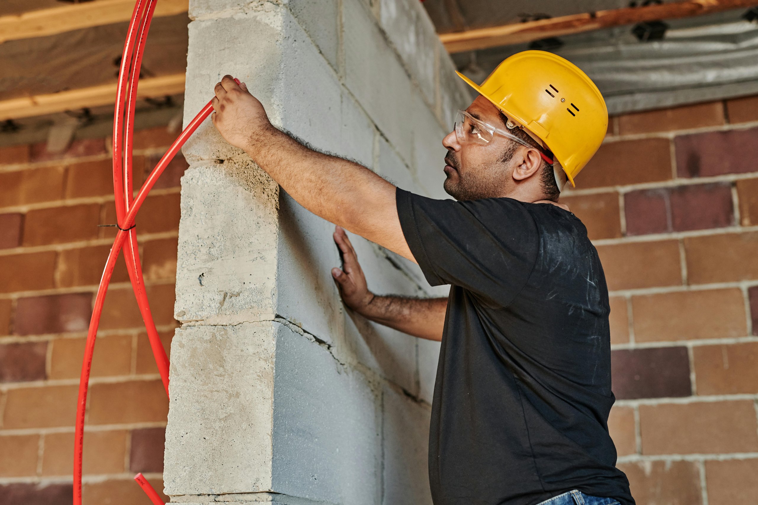 Construction worker in a yellow hard hat installing or inspecting red electrical conduit tubes on a concrete block wall inside a building under construction.