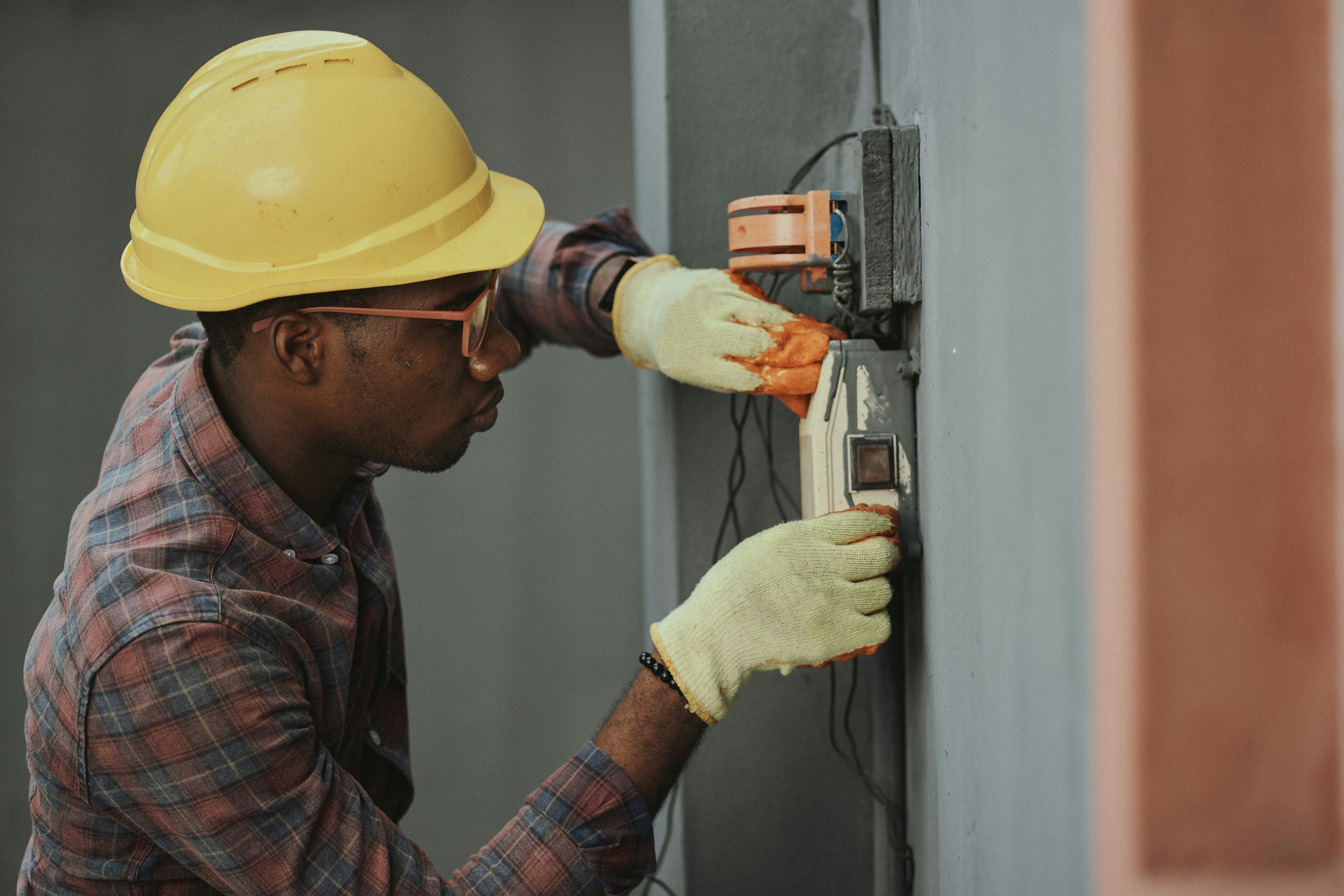 Electrician wearing a yellow hard hat and gloves working on an electrical switch on a gray wall.