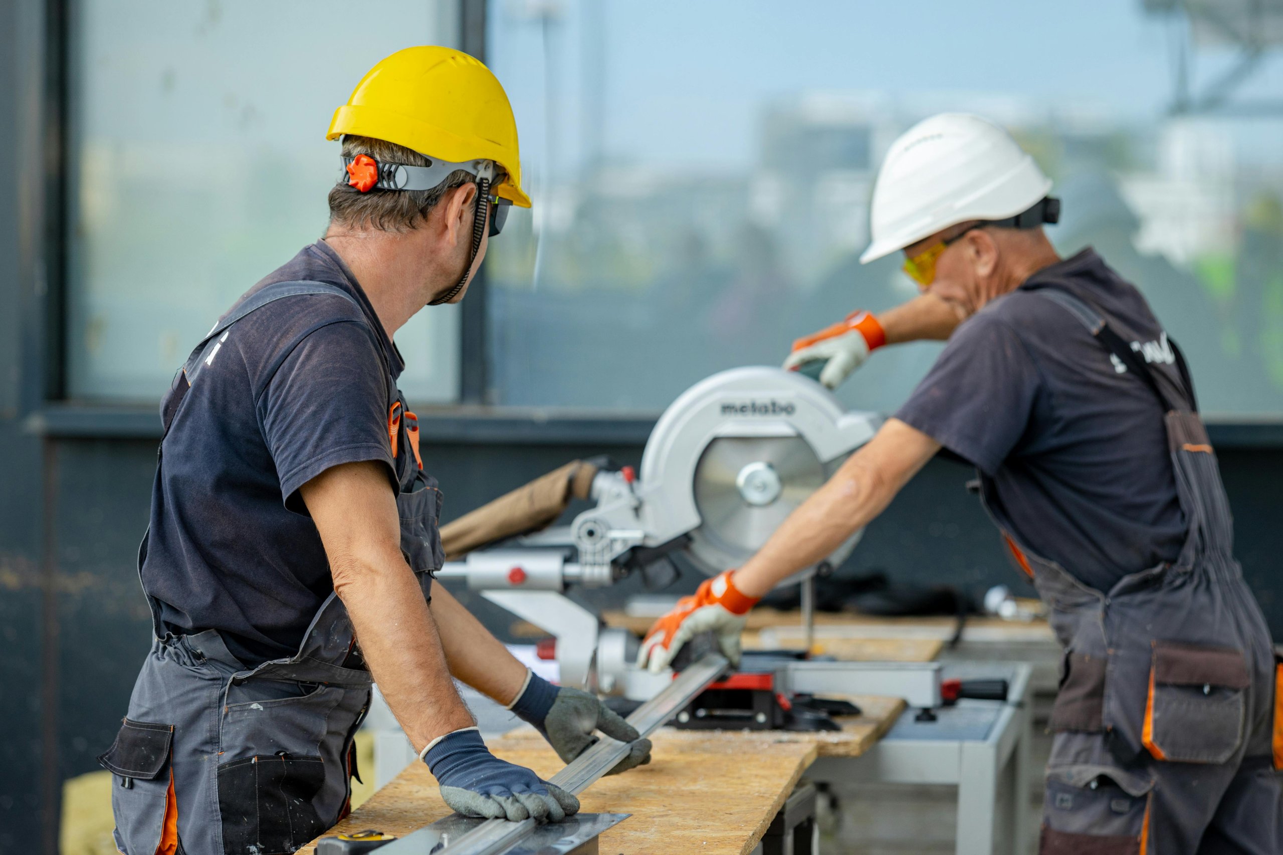 Two construction workers wearing hard hats and gloves operating a miter saw on a wooden workbench outdoors.