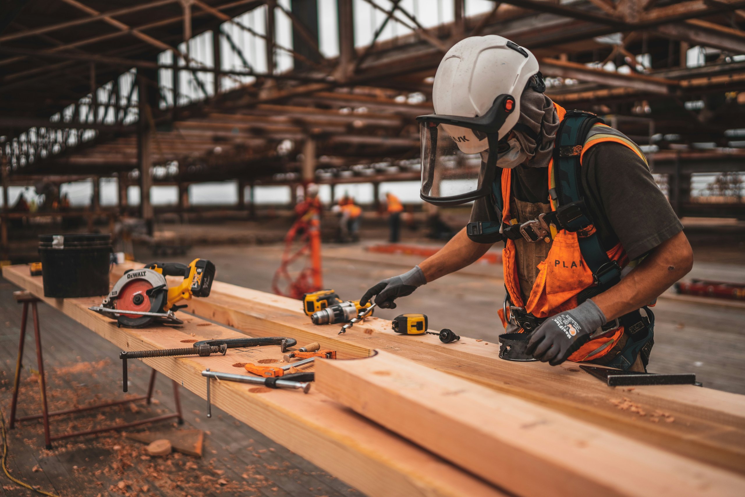 Construction worker wearing protective gear working with tools on a wooden beam inside a large industrial building.