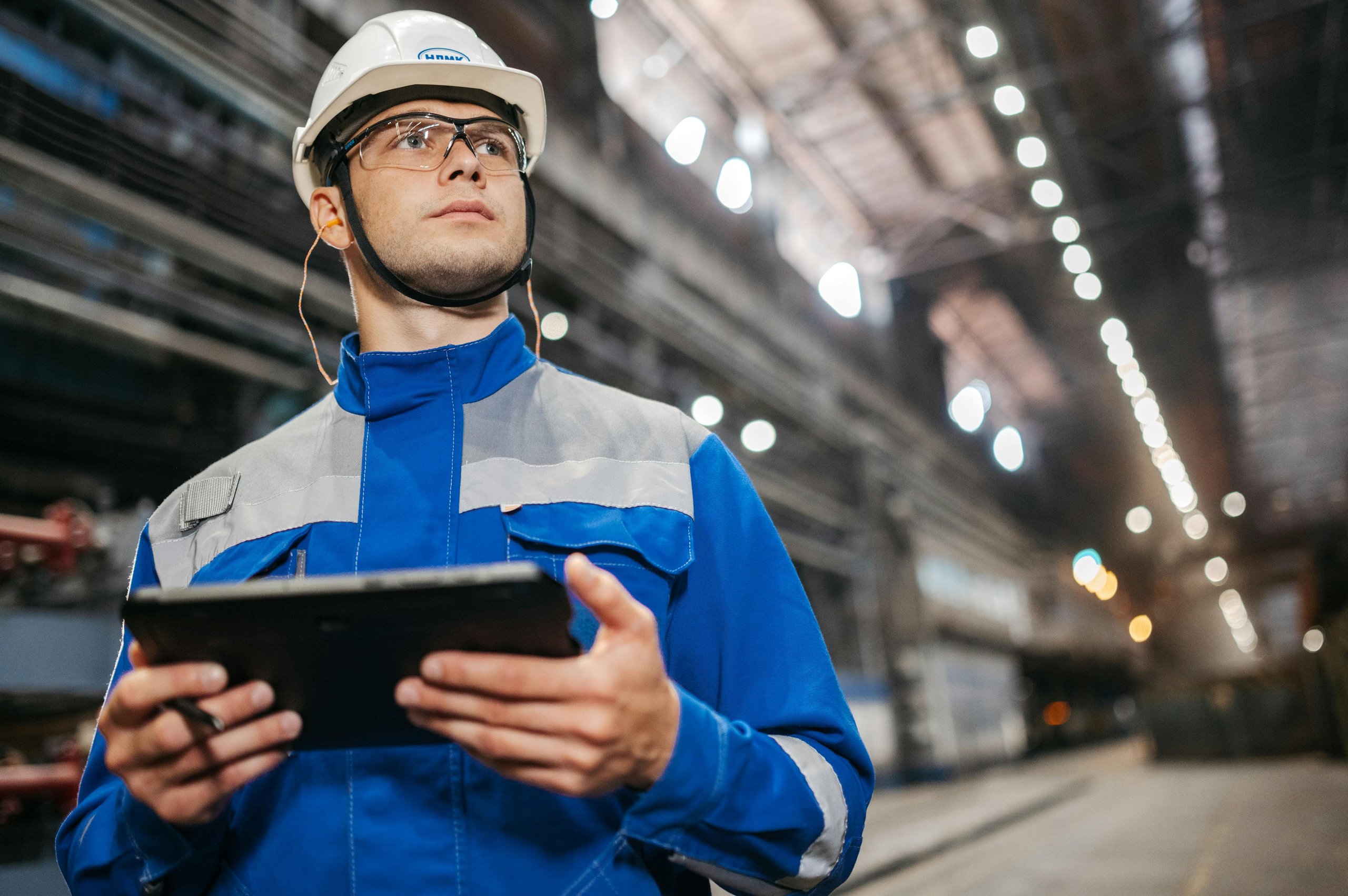 Male factory worker in blue uniform and white hard hat holding a digital tablet inside an industrial facility.