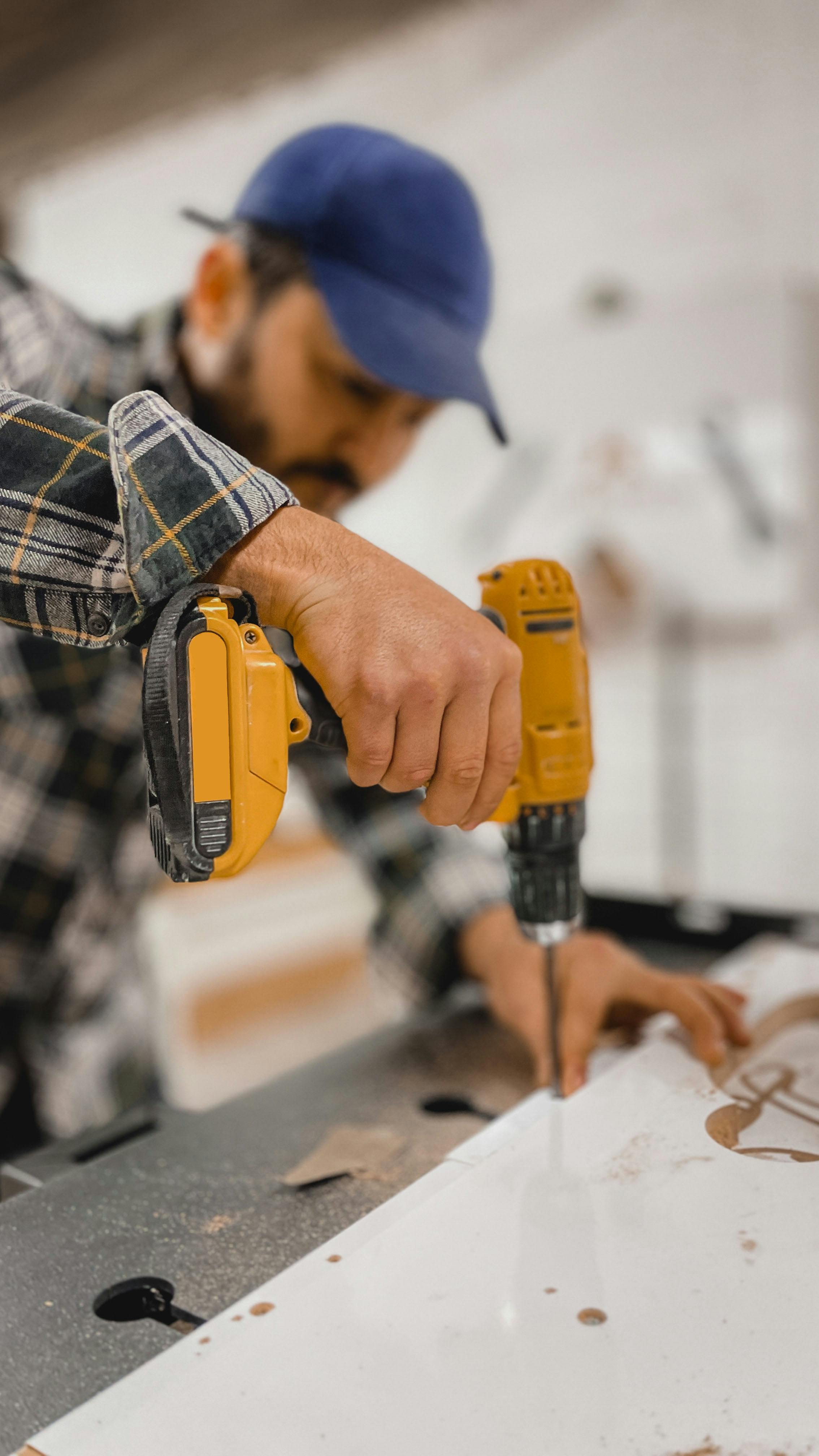 Man in a blue cap and plaid shirt using a yellow cordless drill on a white surface.