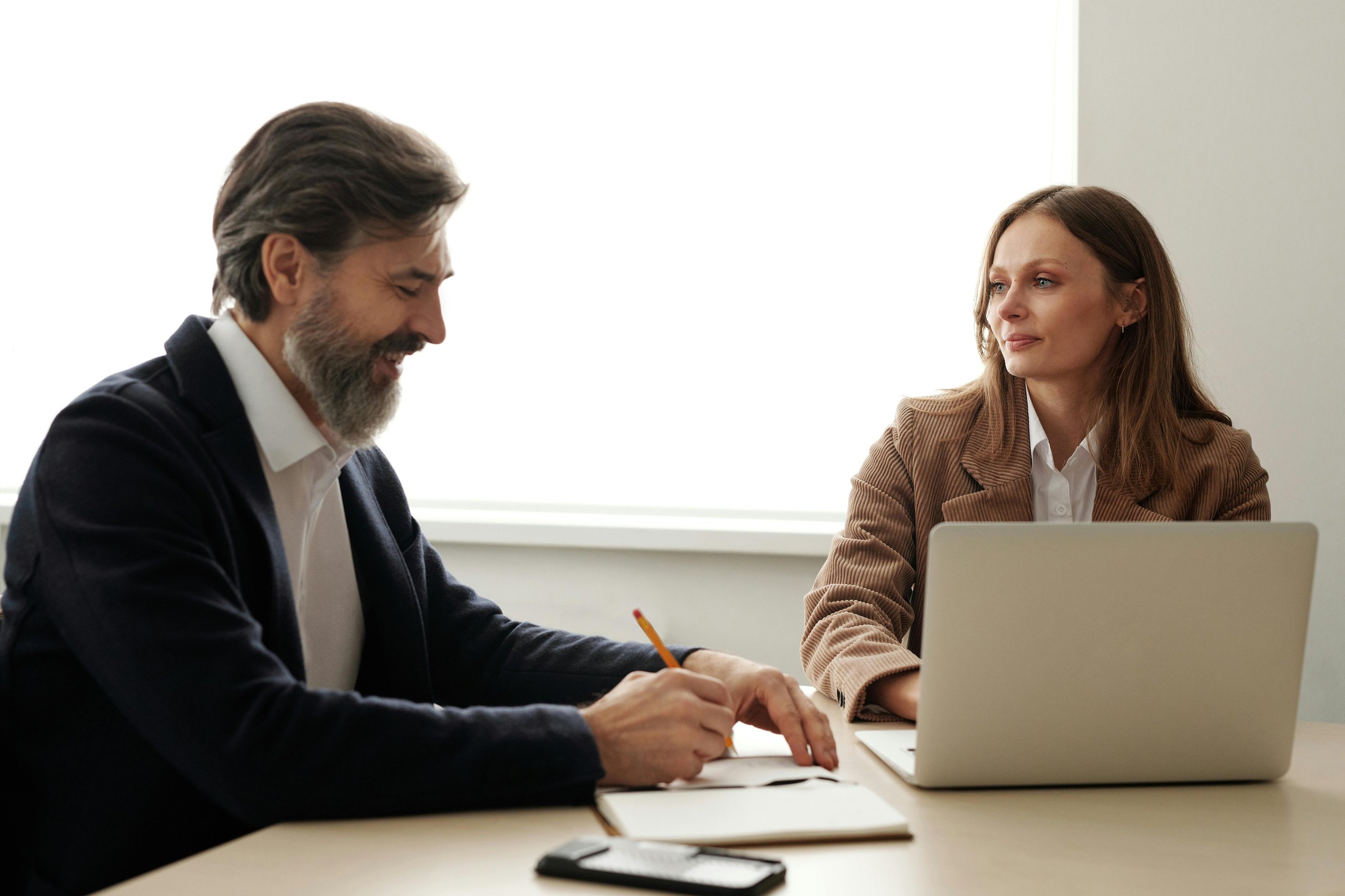 Bearded man writing in a notebook while a woman in a brown jacket looks on during a meeting.