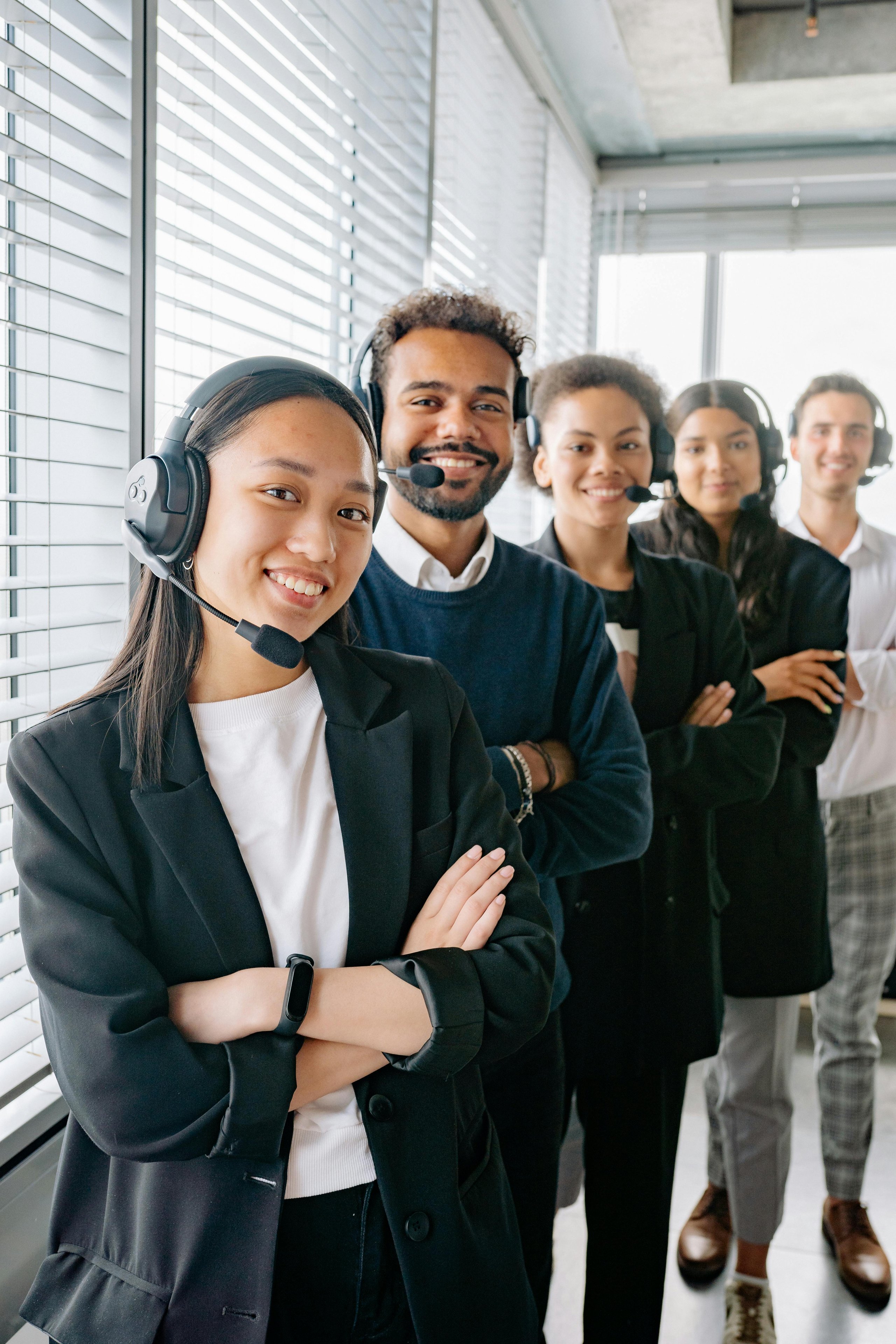 Five diverse customer service representatives wearing headsets standing in a line, smiling with arms crossed.